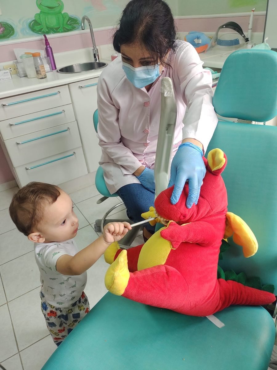 Dra. Marília acompanha um bebê que escova os dentes de um dinossauro de pelúcia sentado na cadeira odontológica.